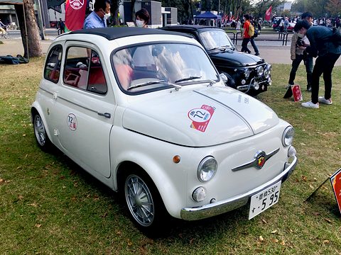 White Fiat 500 with an Abarth moustache badge and a round rally plaque on its bonnet, on grass with visitors behind. Opens a larger view