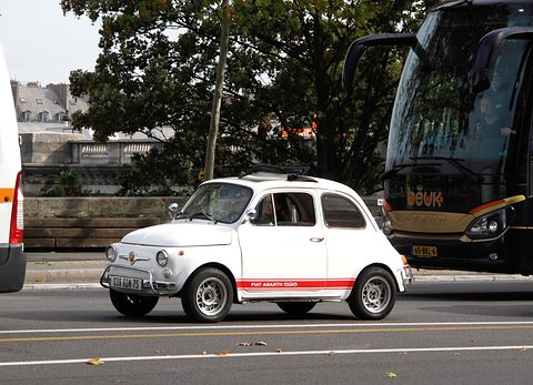 White Fiat 500 with a red waist stripe reading Fiat Abarth 595 on a city road, a black coach behind. Opens a larger view