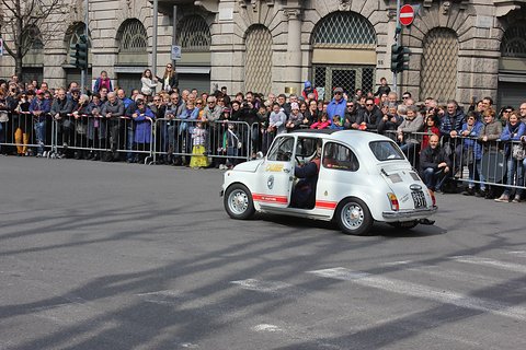 White Fiat 500 with red stripes turning in a city square, a crowd behind barriers and stone buildings beyond. Opens a larger view