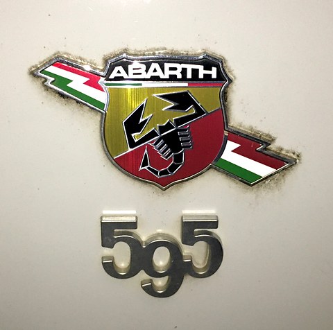 Enamel Abarth shield with a black scorpion and Italian-flag flashes above chrome 595 numerals on cream paint. Opens a larger view