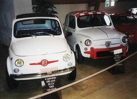 Two white Fiat 500-type cars behind a rope, the left one with a red Abarth moustache badge and a black placard below. Opens a larger view