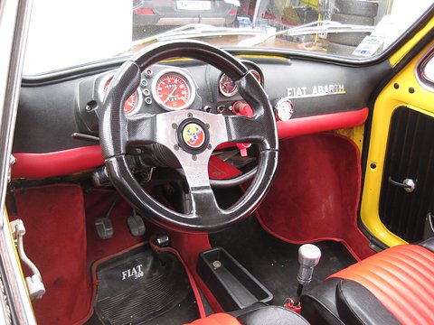 Black three-spoke wheel with an Abarth horn push, red-faced gauges, red carpets and a chrome gear lever.