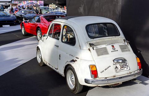 Worn white Fiat 500 with paint chips and Fiat Abarth script on its tail, a red sports car behind. Opens a larger view