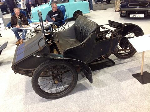 Side view of a black three-wheeled vehicle with a front box, a leather seat and a single rear wheel. Opens a larger view