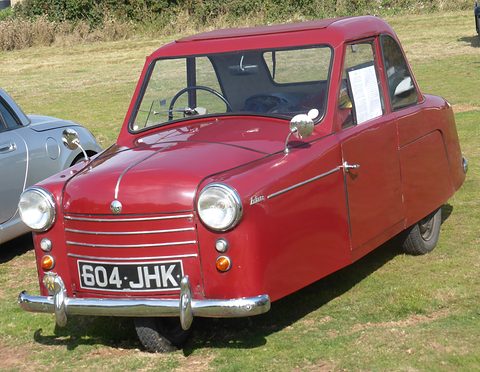 Maroon three-wheel saloon with a chrome-barred grille and two headlamps parked on grass. Opens a larger view