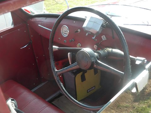 Maroon cockpit with a thin black steering wheel, a small dial and red bench seat. Opens a larger view