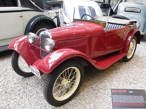 A red open two-seater with cream wire wheels on gravel beside an Aero 10 HP information panel. Opens a larger view