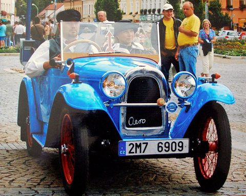 Front view of a bright blue open two-seater with red wire wheels and Aero script on the grille, people around.