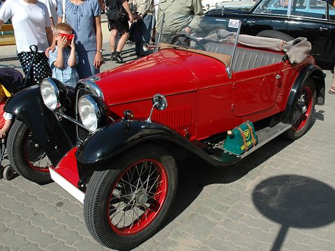 A red open two-seater with black wings and red wire wheels on paving, visitors crowding behind. Opens a larger view