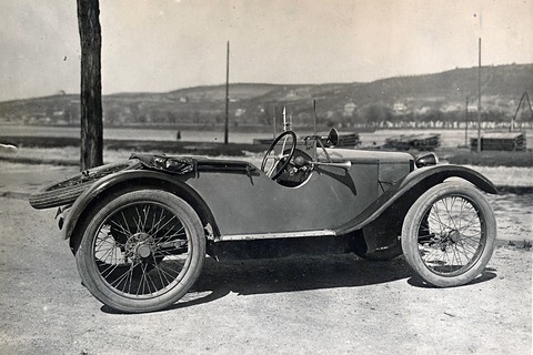 A side view of a small open two-seater with wire wheels, a spare wheel strapped to the tail and no doors. Opens a larger view