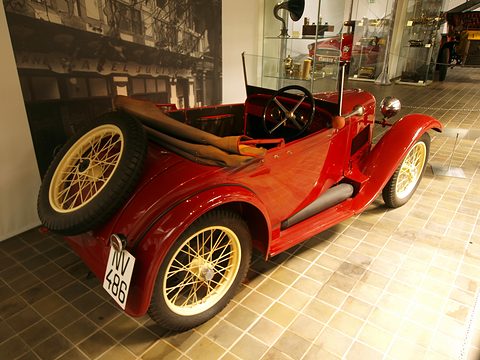 Rear three-quarter view of a red two-seater with cream wire wheels and a spare wheel on its tail. Opens a larger view