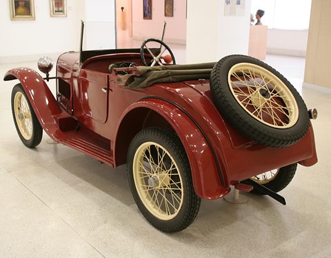 Rear three-quarter view of a maroon two-seater with a spare wire wheel on its tail and a folded hood.