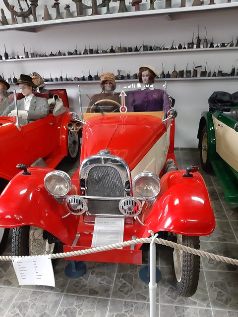 Front view of a red two-seater with a chrome radiator behind a rope, mannequins in period dress behind. Opens a larger view