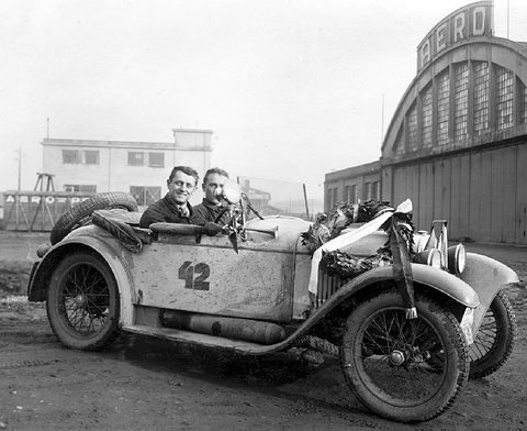 Two smiling men in a mud-spattered open two-seater numbered 42, a wreath on its bonnet, a hangar lettered AERO behind.