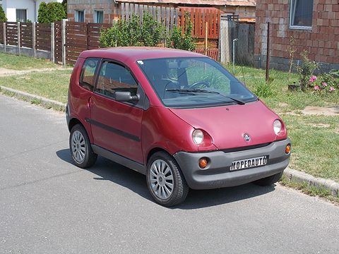 Faded red rounded small hatchback with a MOPEDAUTO plate on its bumper, parked on a quiet residential street. Opens a larger view