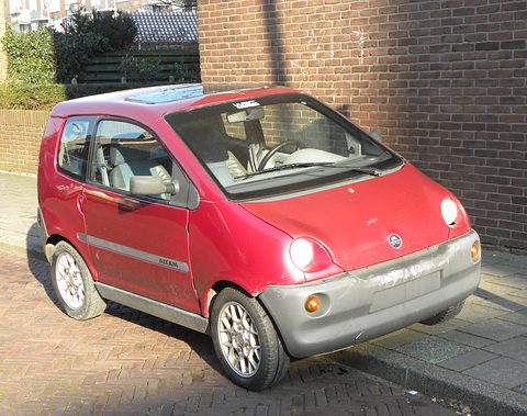 Faded red rounded small hatchback with a grey bumper, parked on brick paving against a brick wall in sunshine. Opens a larger view