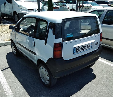 Rear of a white rounded small car with black taped door frames and French plate BE-694-SK, in a sunny car park. Opens a larger view