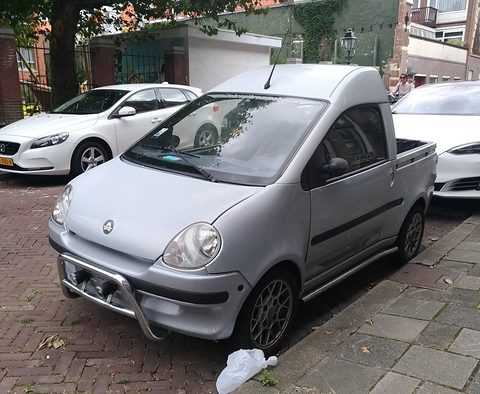 Silver small pick-up with a chrome nudge bar and short load bed, parked on herringbone paving beside white cars. Opens a larger view