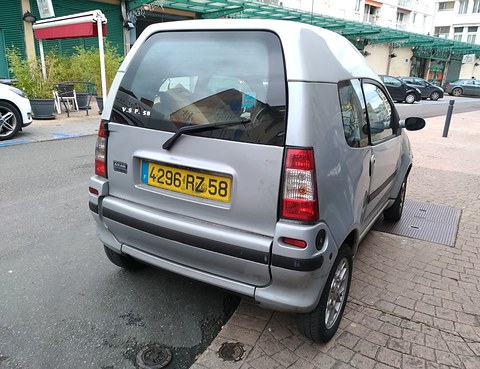 Silver small van-backed car from the rear left, with a large tailgate window and yellow plate, on a paved street. Opens a larger view