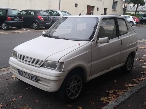 White boxy small hatchback with a slatted grille parked at a kerb strewn with autumn leaves.