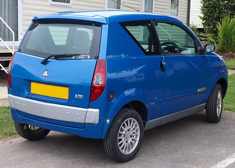Rear of a blue small hatchback with a yellow blank plate and grey bumper, parked by caravans and a palm. Opens a larger view