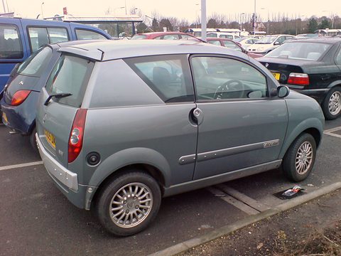 Grey-green small hatchback seen from the rear left in a crowded car park, alloy wheels and a yellow rear plate. Opens a larger view