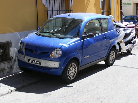 Blue small hatchback with an AIXAM plate, a scooter parked behind it beside an ochre-painted wall. Opens a larger view