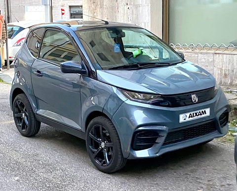 Blue-grey small hatchback with an AIXAM dealer plate and black wheels, a mesh fence and wall behind. Opens a larger view