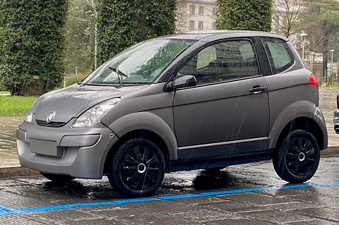 Matt grey small hatchback with black wheels standing on wet paving beside a blue parking line and hedges. Opens a larger view