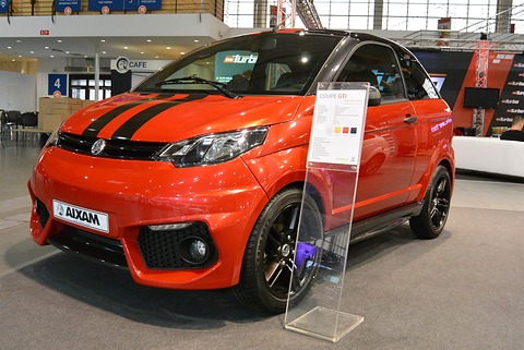 Bright orange small coupé with black bonnet stripes and an AIXAM plate beside a clear acrylic spec stand. Opens a larger view