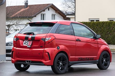 Red small coupé with twin black stripes, black alloy wheels and a 45 speed disc on the tail, parked by a house. Opens a larger view