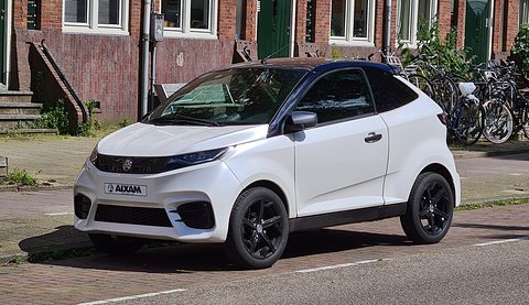 White small coupé with slim headlamps, black grille and an AIXAM front plate, parked by a brick terrace. Opens a larger view