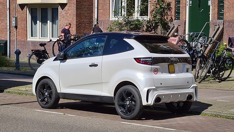 White small coupé with a black roof and black wheels parked by a brick terrace, bicycles and passers-by behind. Opens a larger view