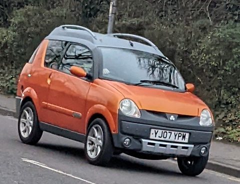 Orange and grey small crossover with roof bars and British plate YJ07 YPM, parked at a kerb before a hedge. Opens a larger view