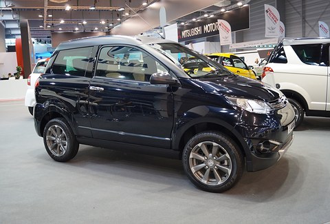 Glossy black small crossover with alloy wheels and chrome-capped mirrors on a grey show floor, a rally car behind. Opens a larger view