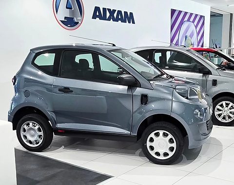 Grey small electric hatchback with plain steel wheels on a white tiled showroom floor, AIXAM lettering on the wall. Opens a larger view