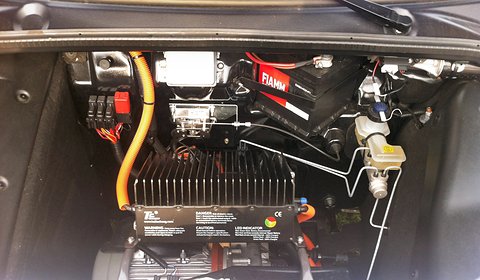 Engine bay with a finned black charger, orange high-voltage cable, a red battery and brake fluid reservoir. Opens a larger view