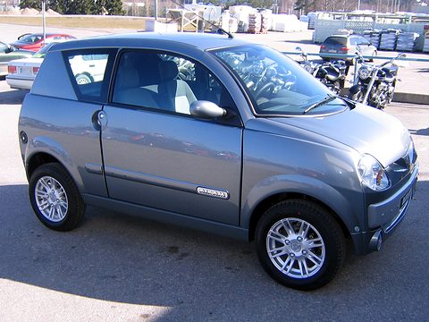 Blue-grey small hatchback side-on in sunshine, motorcycles and pallets of goods behind it. Opens a larger view