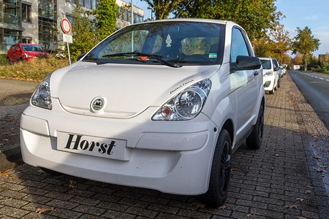 White small hatchback with round headlamps and a novelty name plate on its bumper, parked on brick paving by a road. Opens a larger view