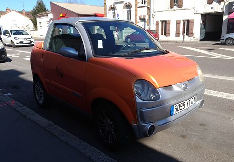 Orange and silver small two-seater with a short open load deck, parked in strong sunshine beside a zebra crossing. Opens a larger view
