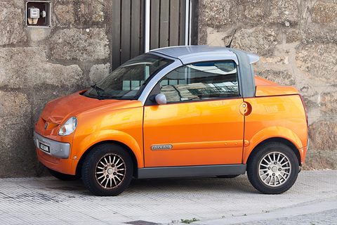 Orange two-seater with a silver roof and short load deck in profile, parked on a pavement before a granite wall.