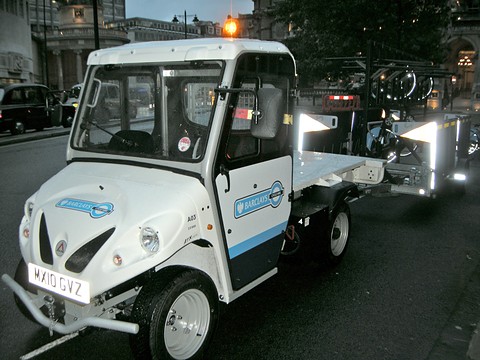 White small electric utility truck with a Barclays logo and blue stripe on a street at night, lights behind. Opens a larger view