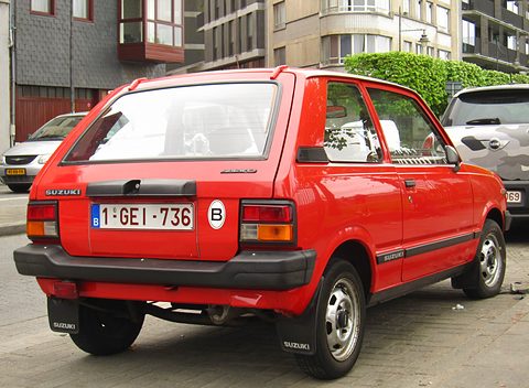 Rear quarter of a red small three-door hatchback with a black rear bumper and a Belgian plate, parked in a street of flats. Opens a larger view