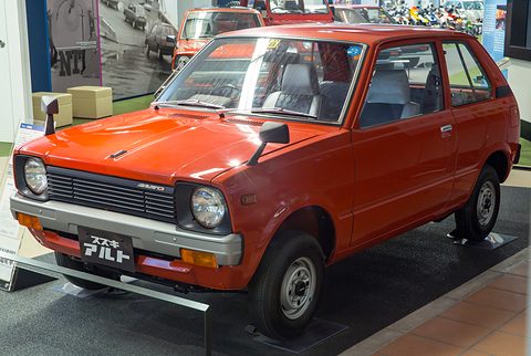 Red small three-door hatchback with round headlamps and a black grille, on a dark plinth in a museum hall.
