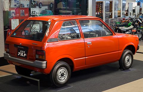 Rear quarter of a red small three-door hatchback with a price card in the rear window, motorcycles on display behind. Opens a larger view