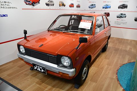 Orange small three-door hatchback with round headlamps and a Japanese show plate, on a wooden floor before a wall of model photographs. Opens a larger view