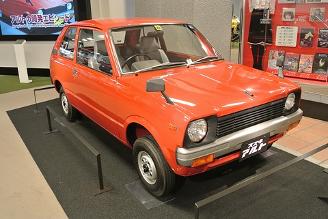 Red small three-door hatchback seen from the front right on a black plinth with low rails, a video screen and posters behind. Opens a larger view