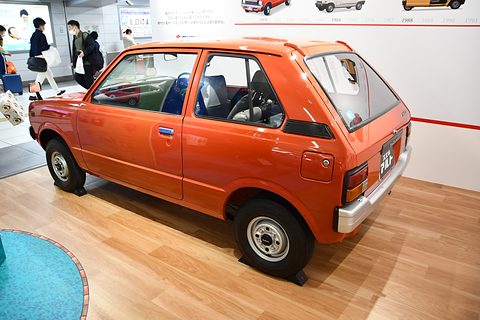 Rear quarter of an orange small three-door hatchback on a wooden floor, a blue round rug and visitors behind. Opens a larger view