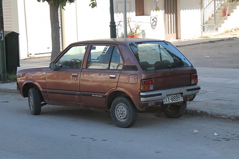 Rear quarter of a brown small five-door hatchback with chrome bumpers, parked on a street by white steps and a tree. Opens a larger view