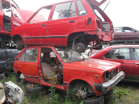 Two red small hatchbacks stacked on top of each other in a scrapyard, bonnets open and parts missing, more wrecks around them. Opens a larger view